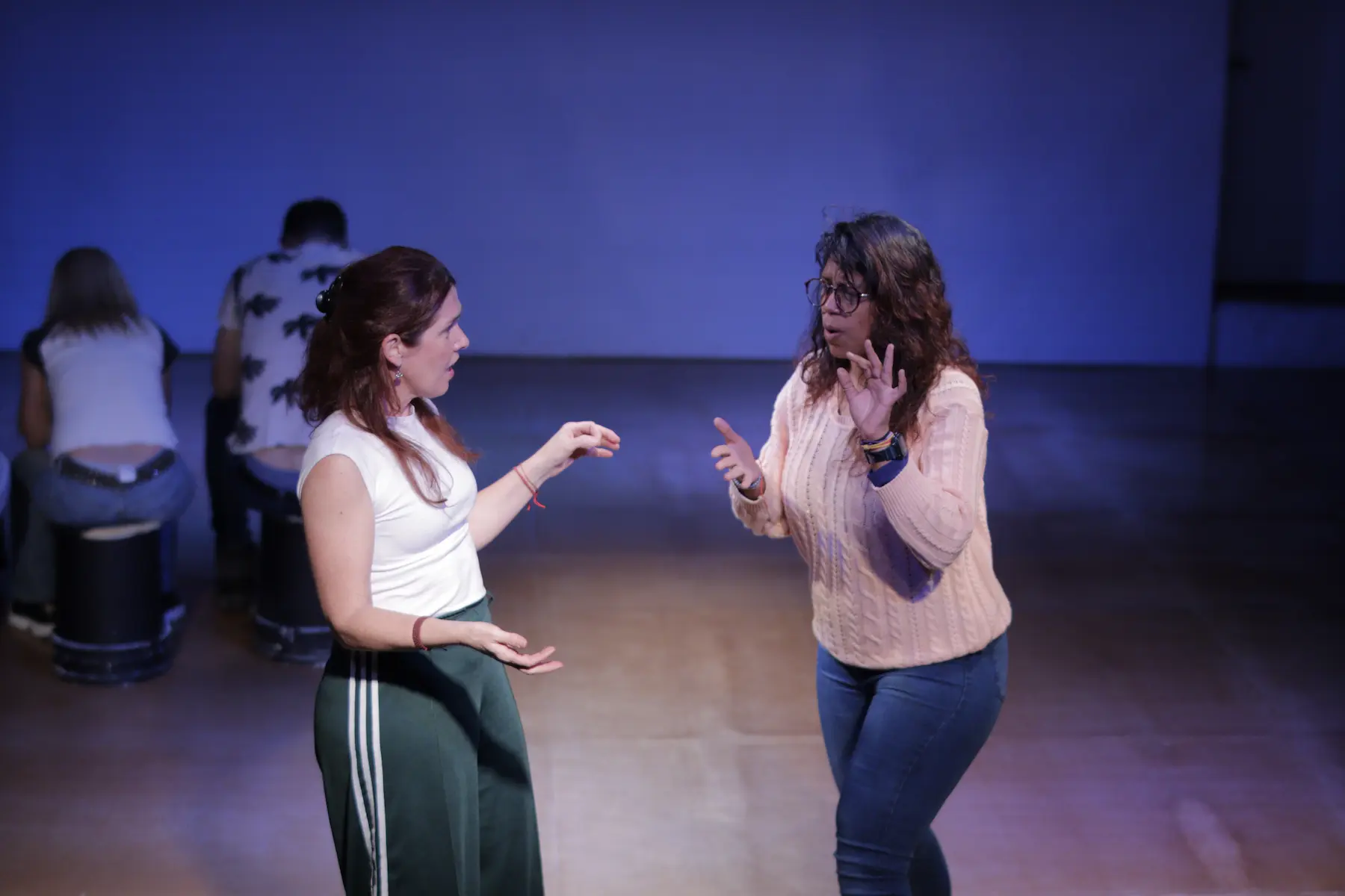 Dos mujeres gesticulando mientras improvisan en una escena, con dos personas de espaldas al fondo